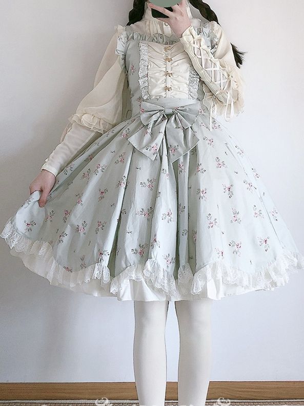 a person is standing indoors wearing a kneelength lolita fashion dress. the dress is light blue with pink floral patterns. it also has lace and ruffles throughout and a large bow at the waist. the person is also wearing a long sleeve white blouse underneath. the background is a white wall with a brown floor.