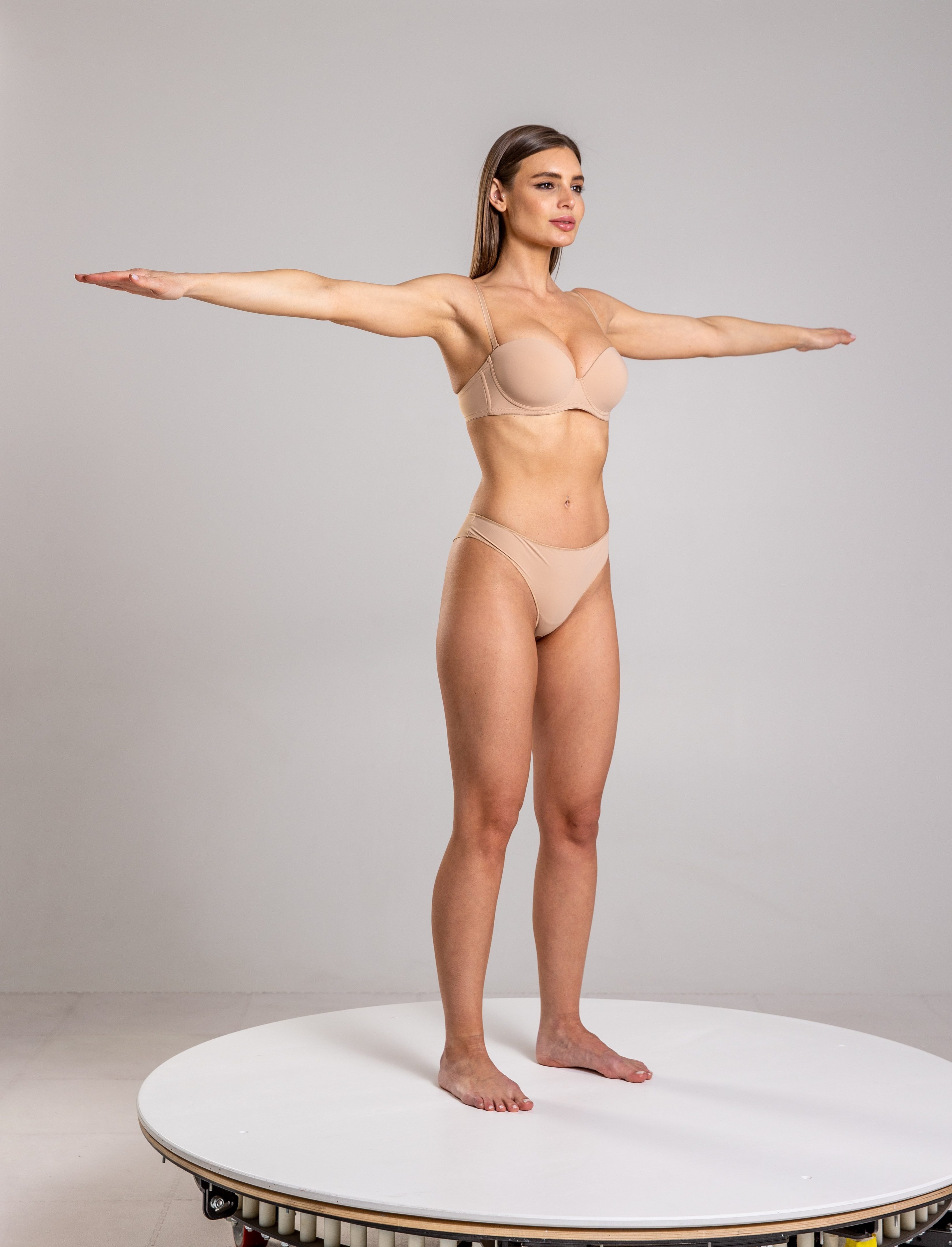 a fullbody studio shot of a woman standing on a white turntable with her arms outstretched. she is wearing neutralcolored underwear and has fair skin. she appears slender and fit. her bare feet are on the turntable. the background is a plain white studio backdrop. the woman is looking to her right with a confident expression.
