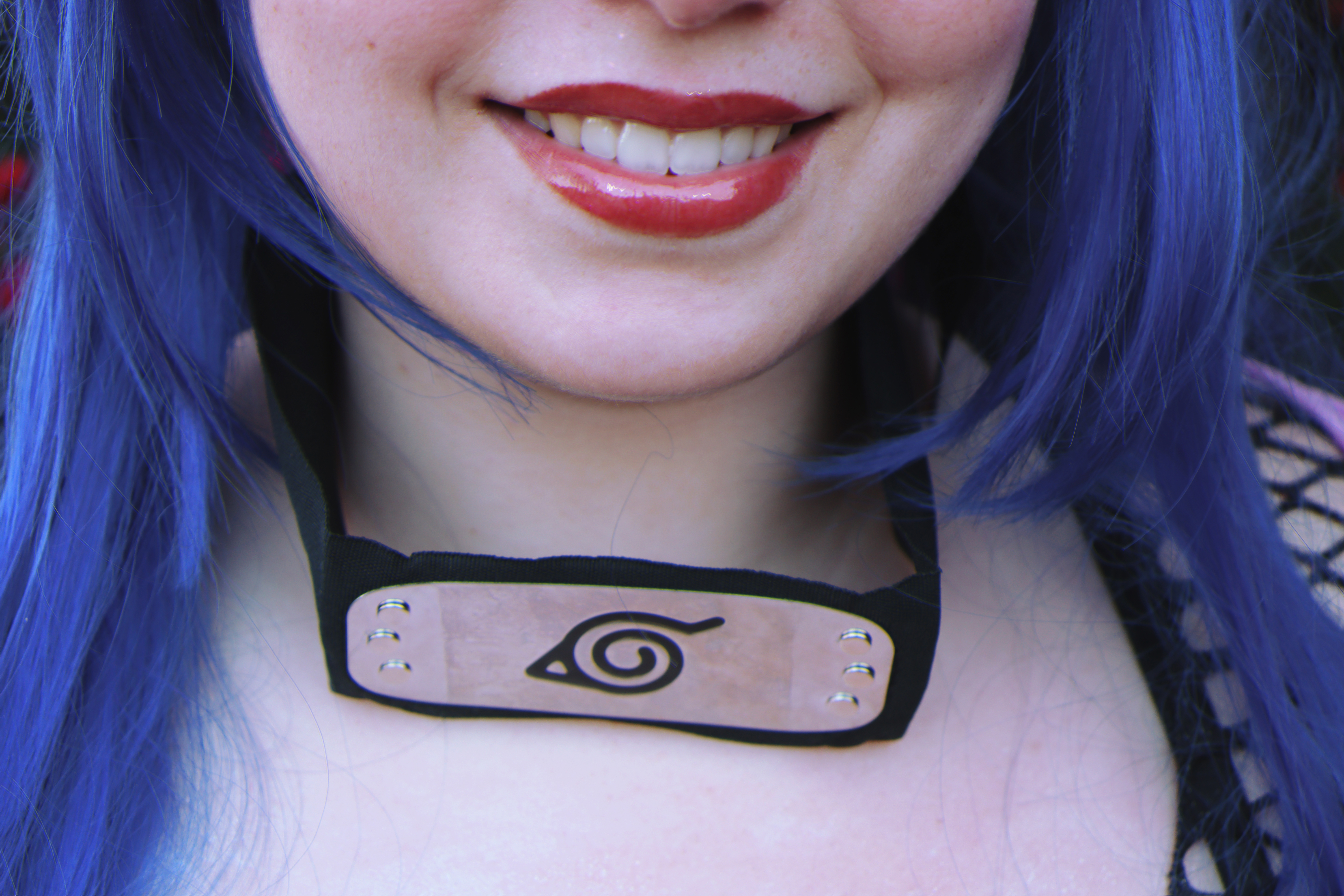 closeup of a person with blue hair, bright red lipstick, and wearing a konoha hidden leaf village headband from the anime naruto. the headband is black with a metal plate in the center displaying the konoha symbol. the person is smiling. the image suggests a konan naruto cosplay.
