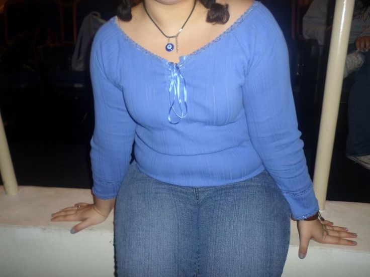 a woman is sitting down, wearing a blue shirt and jeans. she has a necklace on and her hair is in pigtails. her hands are resting on the surface she's sitting on.