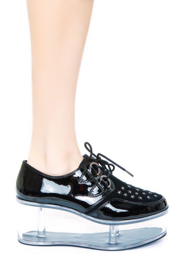 a person's leg and foot are shown wearing a shiny black shoe with laces and small stars. the shoe features a clear platform sole with transparent supports. the background is white.