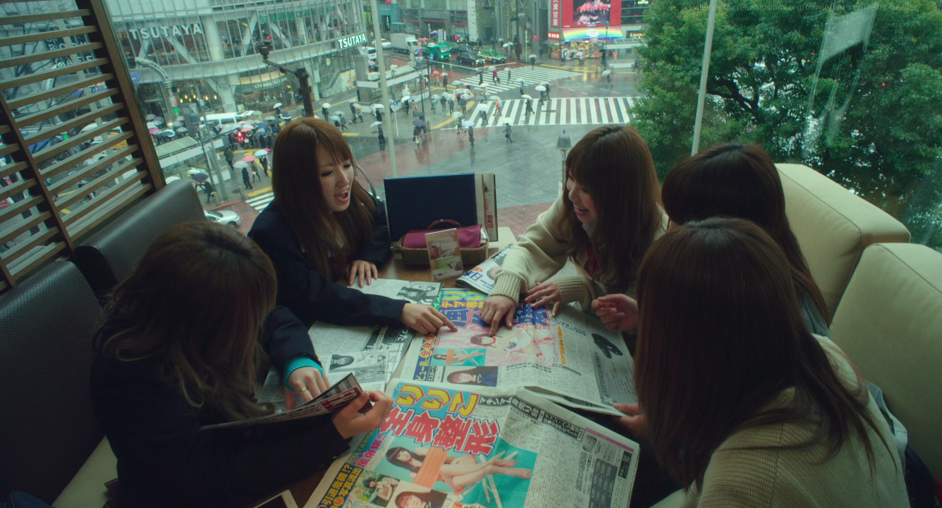 a group of young women are gathered around a table in a cafe, looking at magazines and talking. the cafe has large windows with a view of a busy city street and crosswalk. the day is overcast and the lighting is soft.  the overall tone of the image is casual and friendly.