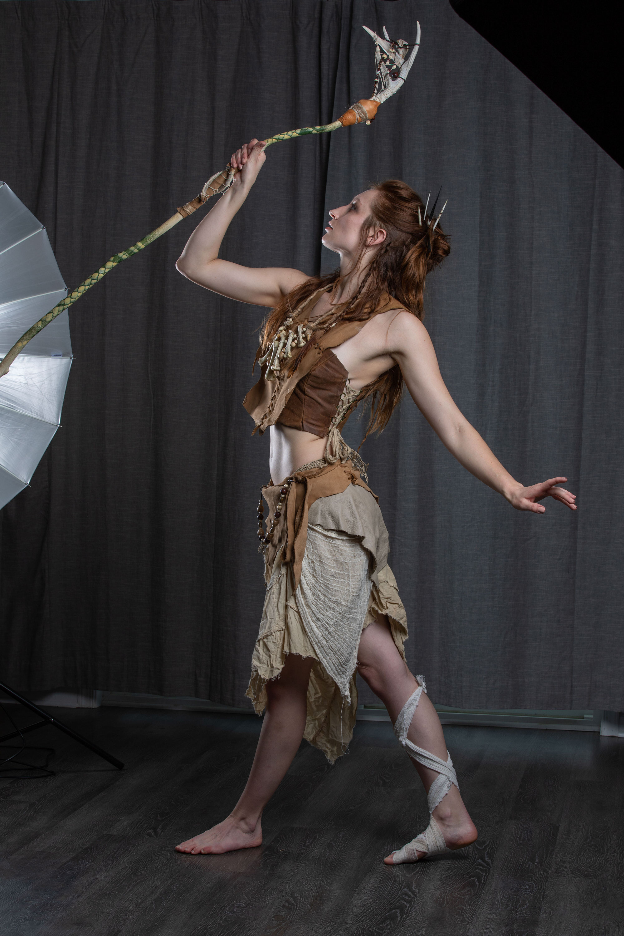 a woman in a tribalinspired costume is posing in a studio. she is holding a staff with a bone ornament on top of it. the background is dark and the lighting is artificial.  she is looking up and to her right as she balances on one foot.