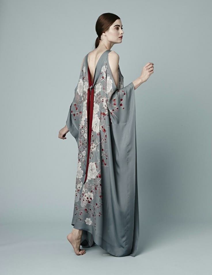 a woman stands in a studio wearing a long gray kimono dress with a floral print. the dress has a red panel down the back and wide sleeves. the woman has a ponytail and is barefoot. she is turned to the side, looking away from the camera.