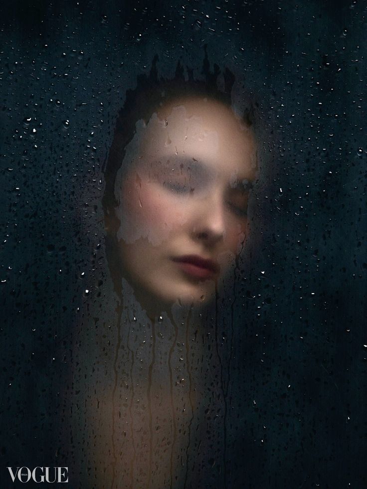 the image is a closeup portrait of a woman's face seen through a misty or raincovered glass. her face is partially obscured by the water droplets, creating a dreamy, ethereal effect. the overall mood is dark, mysterious, and sensual. the vogue logo is visible in the bottom left corner, suggesting the image is from a fashion magazine. the focus is soft, enhancing the dreamy atmosphere. the woman's eyes are closed, and her lips are painted a dark shade.  the background is a dark, blurry expanse. the overall composition is artistic and evocative.