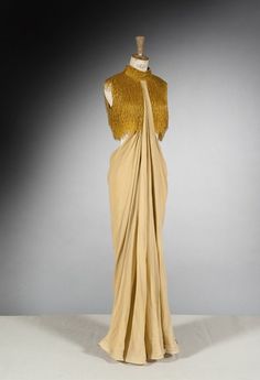 a vintage fashion dress on a mannequin. the dress is gold and has a very classic, elegant style to it. the dress is a formal gown that would be worn to an upscale event. the dress is photographed in a studio setting, with the focus on the dress itself. the dress is long and flowing. the dress is made of a beautiful material that is silky and smooth. the dress is a beautiful piece of vintage clothing.