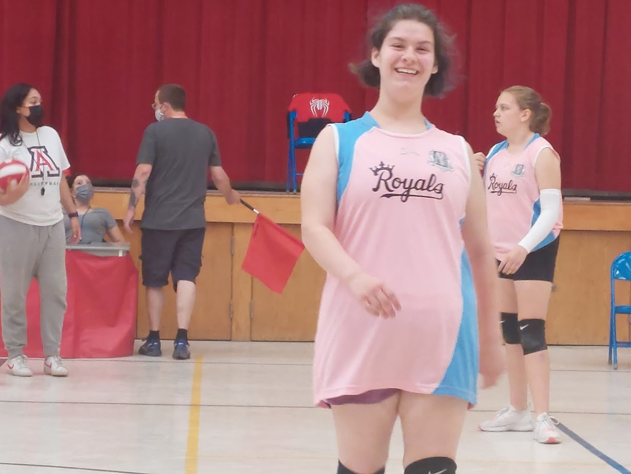 four females are in a brightly lit gymnasium. the female on the far left is holding a volleyball and appears to be the coach. the three other females are wearing matching pink and blue volleyball uniforms and are players on the same team. the player in the middle is smiling and appears to be enjoying herself.  based on the attire of the people in the gym and the red boundary line behind the players, it appears that they are practicing volleyball.