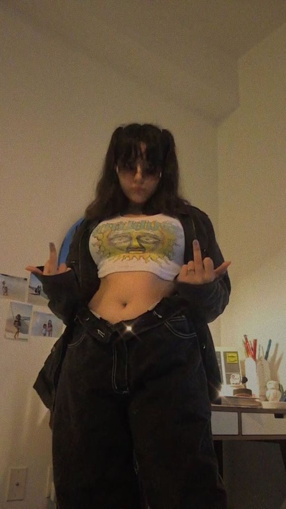 a young woman is taking a mirror selfie in her bedroom. she is wearing a white cropped graphic tee, black jacket, and lowrise black jeans. her hair is styled with two pigtails and bangs.  the room is dimly lit with artificial light and decorated with personal items like photos and art supplies.
