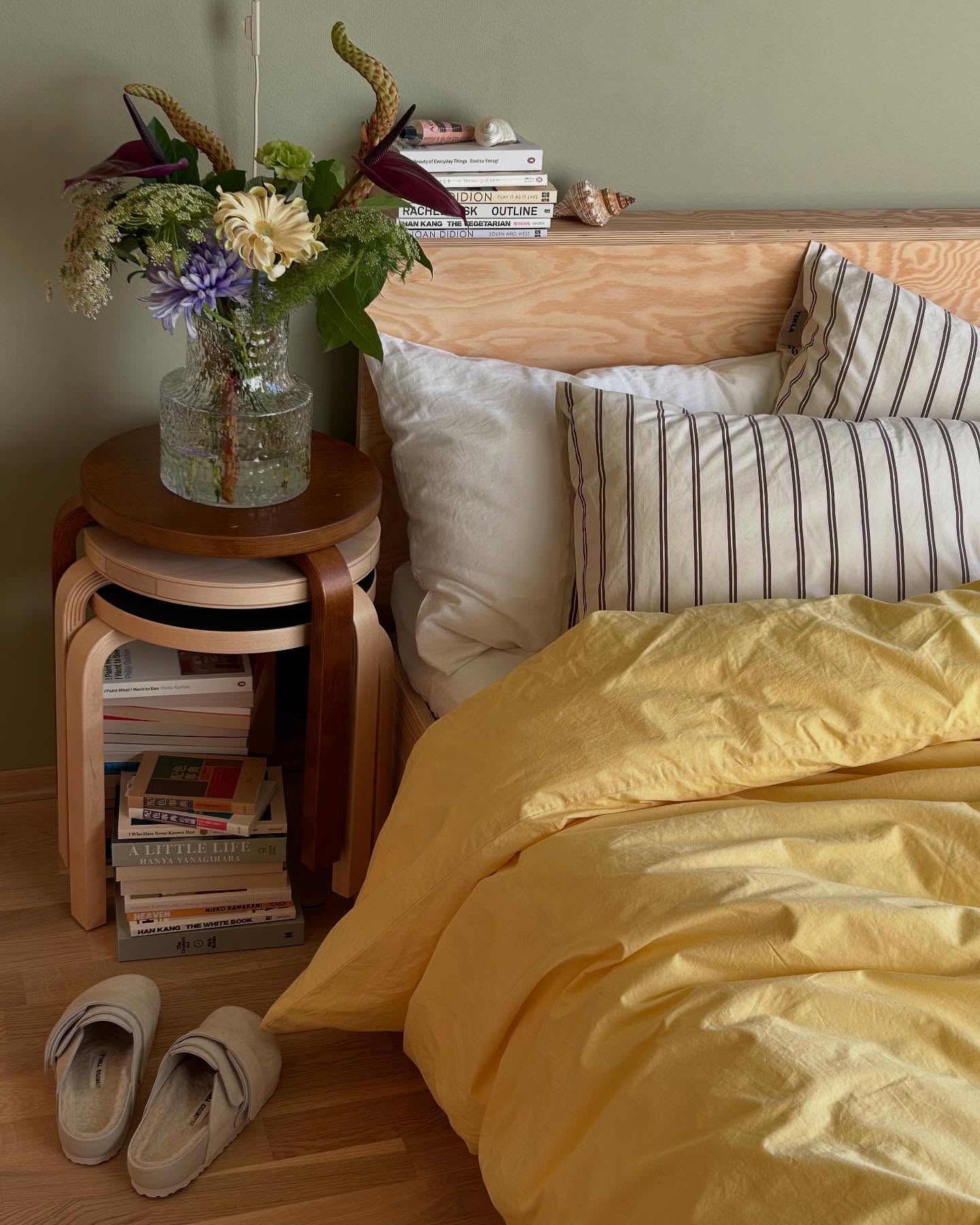 a cozy bedroom scene with a bed adorned with a yellow duvet and various pillows against a wooden headboard. a side table made of stacked stools holds a vase of flowers and a stack of books. slippers are placed on the wooden floor beside the bed. the room is decorated with a natural, homey aesthetic.