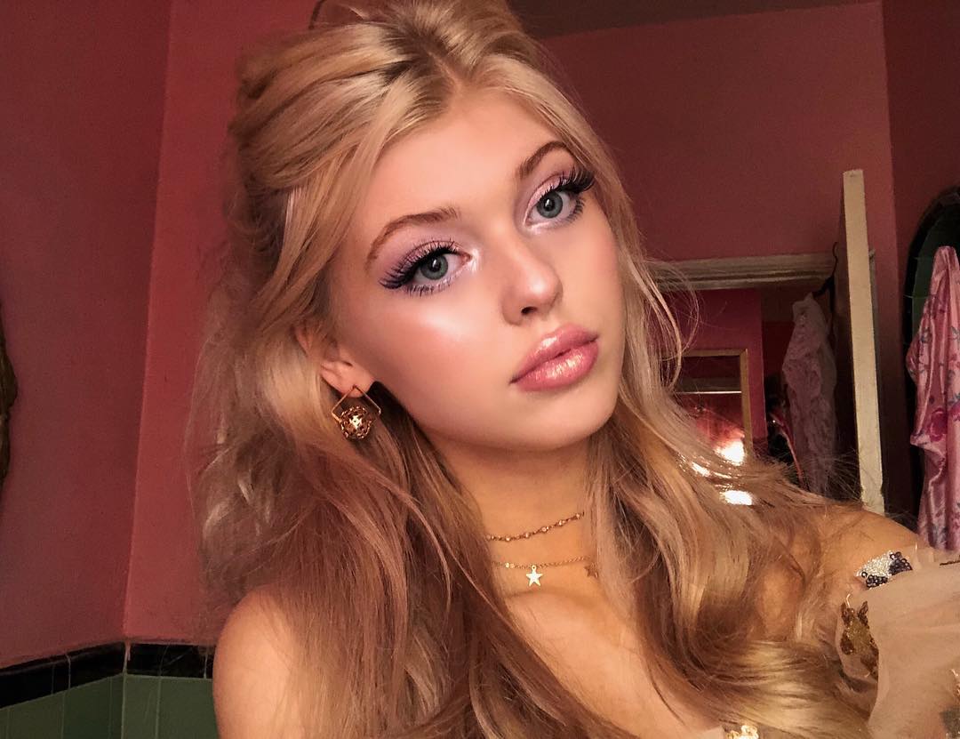 a portrait of a blonde woman with pink eyeshadow, long lashes, gold earrings, and a star necklace, standing in a pink bathroom.