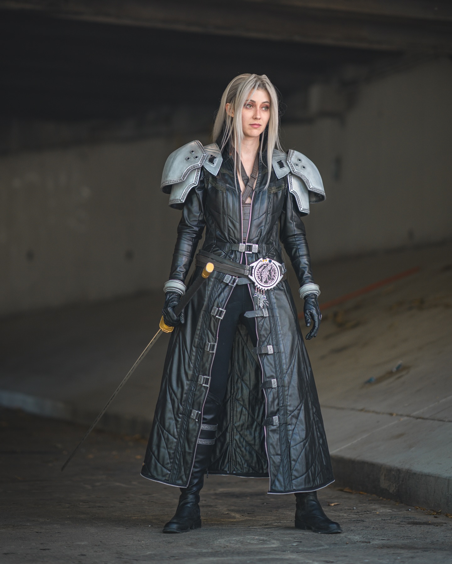 the image is a fullbody shot of a woman cosplaying as a female version of sephiroth from the video game final fantasy vii. she is standing in a dark, urban underpass.  her cosplay is highly detailed, featuring a long black leather coat with shoulder pauldrons, black pants, and tall boots. she's wielding a sword and her long, grey hair is styled to resemble sephiroth's. the setting and costume create a dramatic and visually appealing image. the focus is sharp, and the lighting effectively highlights the costume and the character's pose.