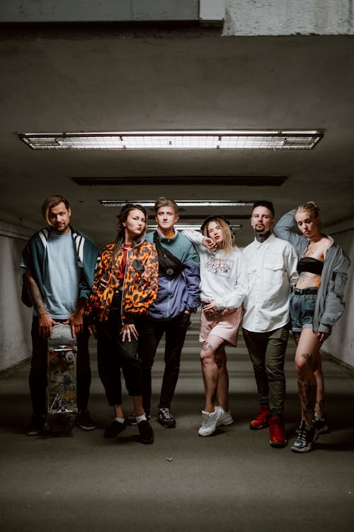 a group of young adults, possibly a band, stand in a dimly lit urban tunnel.  the group is diverse, with both male and female members, and their style is casual and alternative.  one person holds a skateboard. they appear to be posing for a photograph. the atmosphere is cool and edgy.