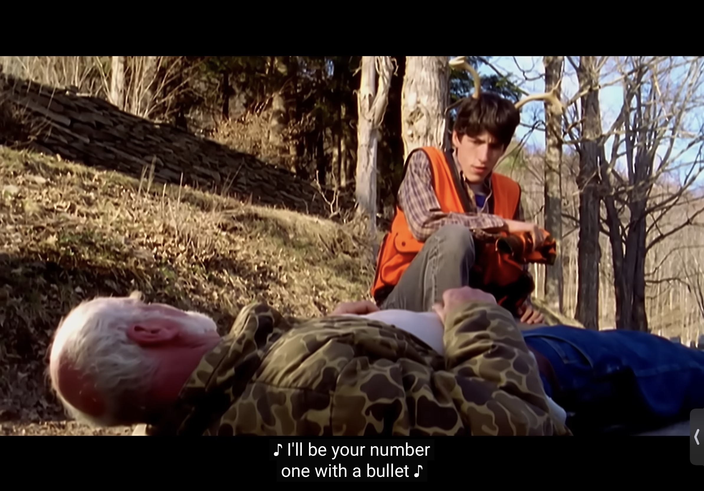 a young man in an orange hunting vest is sitting with a rifle in his hand. he is looking down at an older man who is lying on the ground, presumably injured. the scene takes place in a woodland setting, with trees and a stone wall in the background. the man is wearing a camouflage jacket. this image likely depicts a hunting accident.