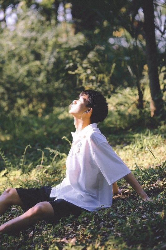 a young asian man, wearing a white shirt and black shorts, is sitting on a grassy slope in a forest. he has his eyes closed and his head tilted back, appearing to be enjoying the dappled sunlight filtering through the trees. the scene evokes a sense of peace and relaxation.