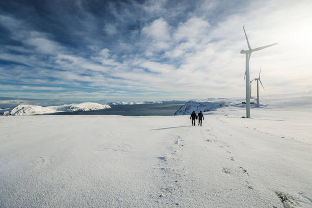 two people walk in a snowy, arctic landscape with wind turbines in the distance. the mountains in the distance and the vast expanse of white snow create a beautiful scene.