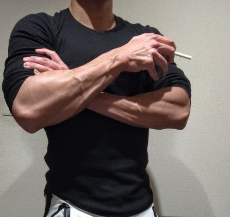 closeup view of a muscular man with prominent veins in his arms. he's wearing a black, closefitting longsleeved shirt. his arms are crossed over his chest, and he's holding a cigarette between his fingers. the background is a plain, light beige wall. the image emphasizes the subject's physique and vascularity. the overall mood is neutral.