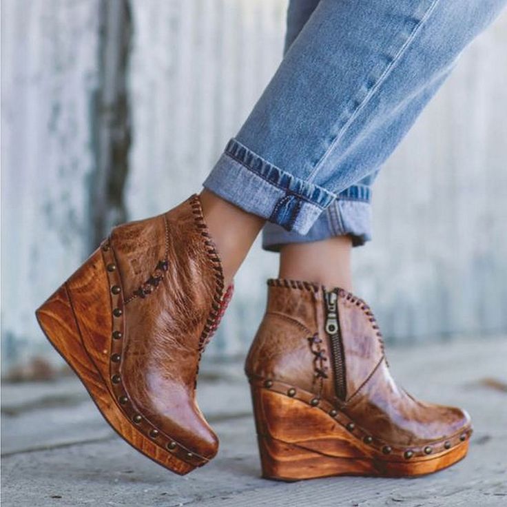 a closeup shot of a woman's feet wearing a pair of brown leather wedge ankle boots. the boots are distressed and have wooden wedges with visible studs. the woman is wearing cuffed jeans and the setting appears to be outdoors. the image showcases the boots' details and style, suggesting a bohochic or western aesthetic.