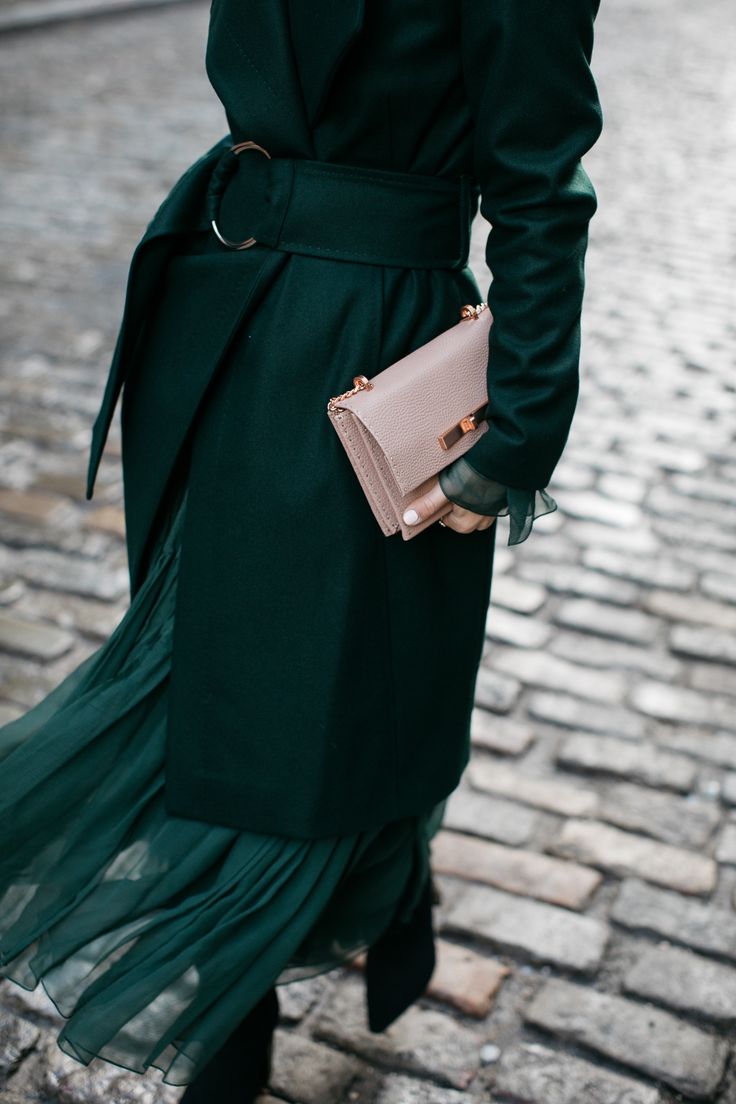 a woman in an emerald green coat and pleated skirt walks down a cobblestone street. she carries a light beige handbag. her style is elegant and sophisticated. the photo showcases fall/winter fashion.
