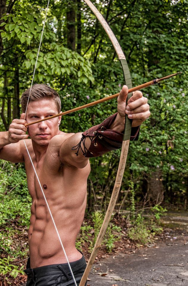 a man in the woods, shirtless and holding a bow and arrow in his hand, aiming at a target.  he is surrounded by nature and green trees.