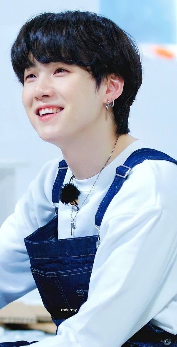 a person with dark hair wearing blue denim overalls over a white longsleeve shirt is smiling and looking away from the camera. they are wearing silver hoop earrings and a silver necklace with a fluffy ball charm. the person is identified as a korean kpop idol and singer. the bottom of the image contains the text "mdarmy".