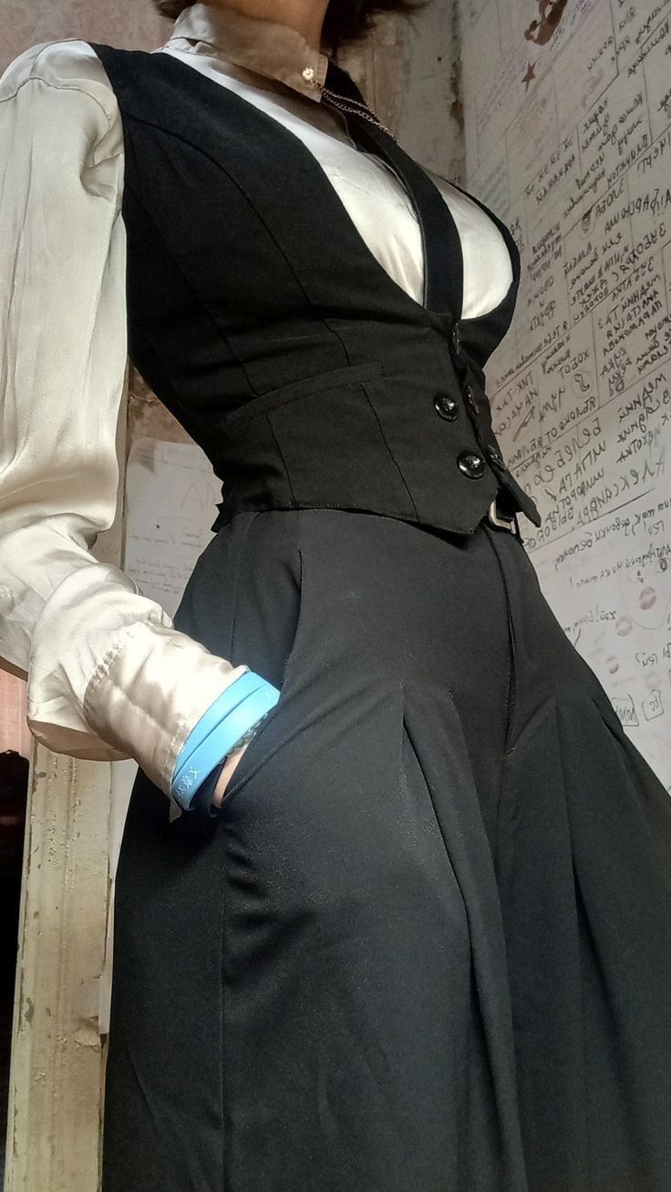 a woman is wearing a white blouse with a black vest and black pants. her hand is in her pocket. she is standing indoors in front of a wall with writing on it.