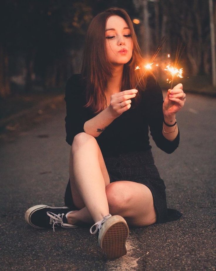a young woman is sitting on the street at night, holding two lit sparklers. she is looking at them with a serene expression on her face. the lighting is low key and warm, creating a magical and celebratory atmosphere.