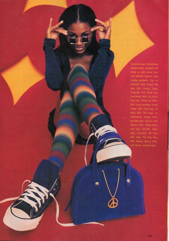 a young black woman model wearing colorful striped socks, hightop converse sneakers and a blue handbag with a peace sign charm. she is posing in a sitting position with her legs crossed and is holding her sunglasses. she is sitting in front of a red and yellow background. the image looks to be from the 1990s.