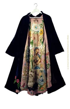 a black cape with an embroidered pink dress underneath. the dress has flowers and a religious scene in the center. the cape is gathered at the neck and has a velvetlike texture.  the clothing appears to be historical, possibly from the victorian era.  the scene on the dress is religious, likely depicting a saint or holy figure.  the style is formal and elegant.
