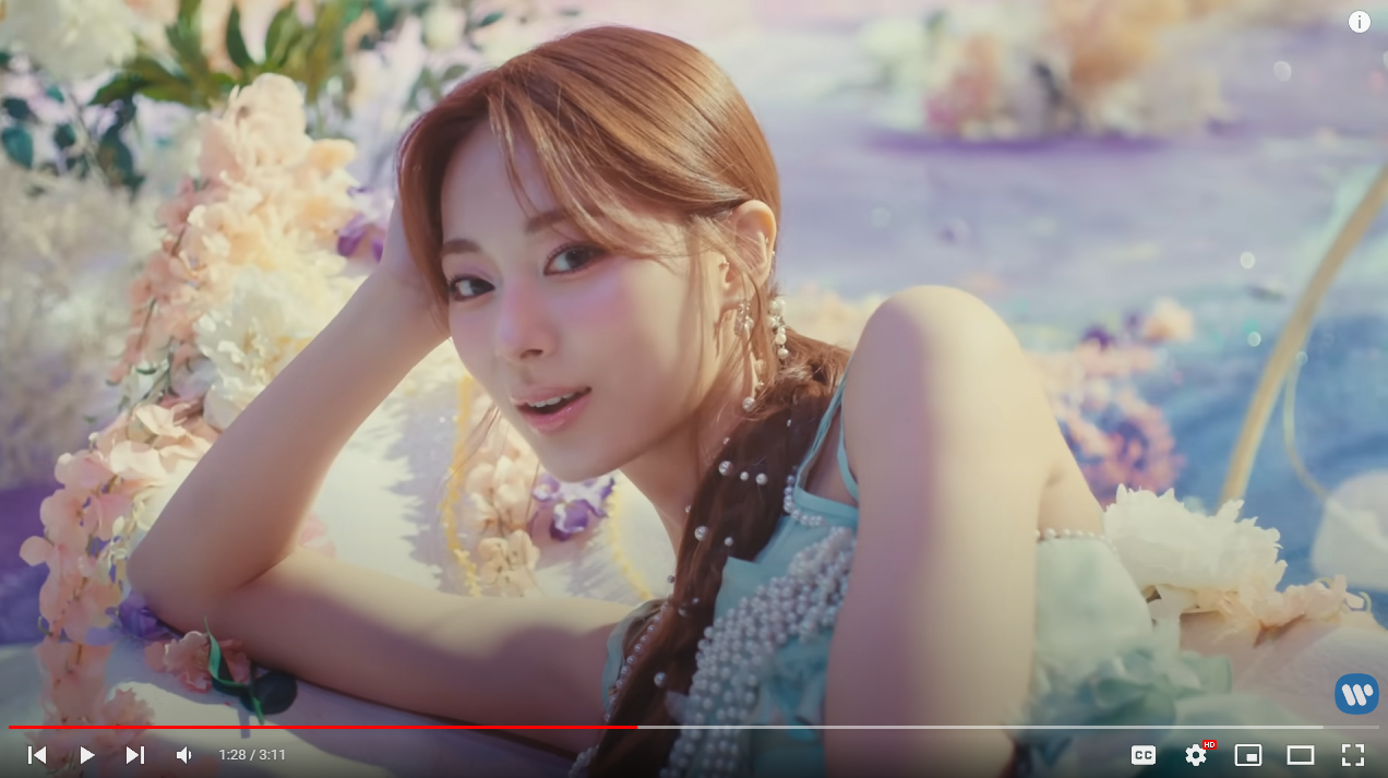 a screenshot of jeongyeon from the kpop group twice, likely from a music video. she is laying down in a pastel dreamlike setting, with soft lighting. she is wearing a light blue top adorned with pearls and a halfup ponytail hairstyle. the scene exudes a soft and feminine aesthetic.