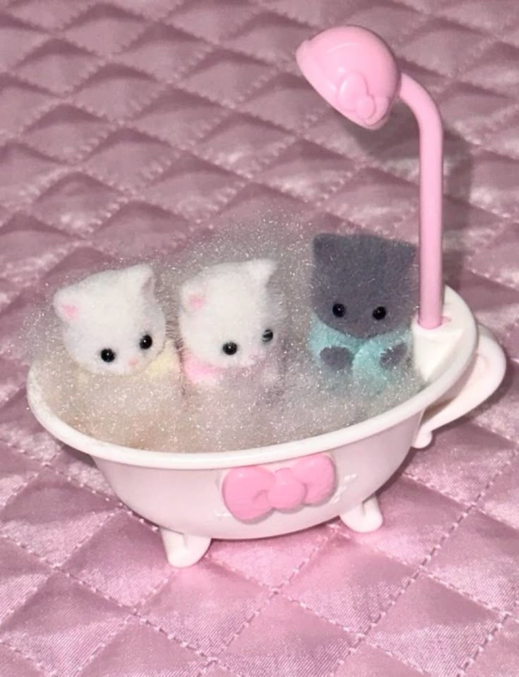 a photo of three calico critters kittens in a pink bathtub filled with bubbles. the bathtub has a pink showerhead attachment and a pink bow on the front. the kittens are one gray and two white, and they look like they are enjoying their bath. the scene is set on a pink quilted surface.