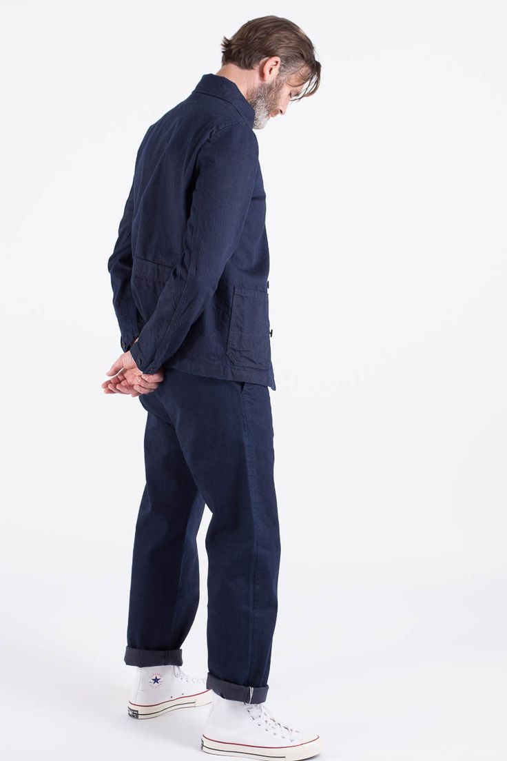 a man is standing in a white studio. he is wearing a dark blue jacket and pants. he is also wearing white converse shoes. he has a casual fashion style.