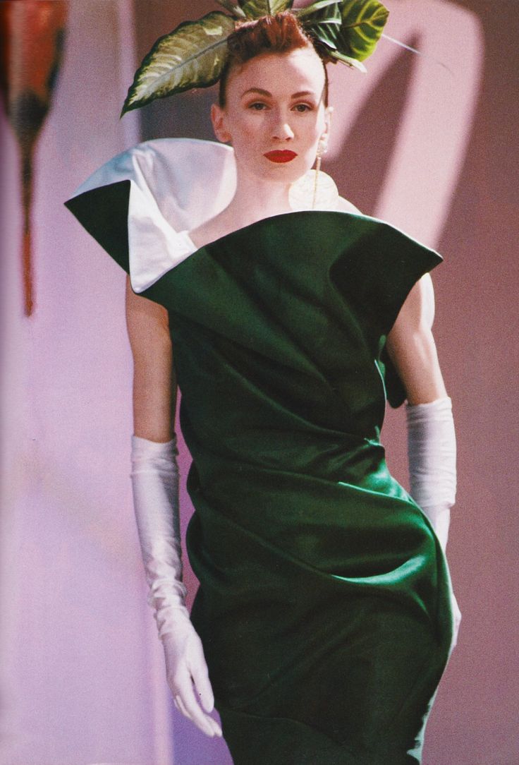 a woman is posing in a green dress with white trim, white gloves, and large green leaves in her hair. she is wearing red lipstick. the background is a muted pink color.