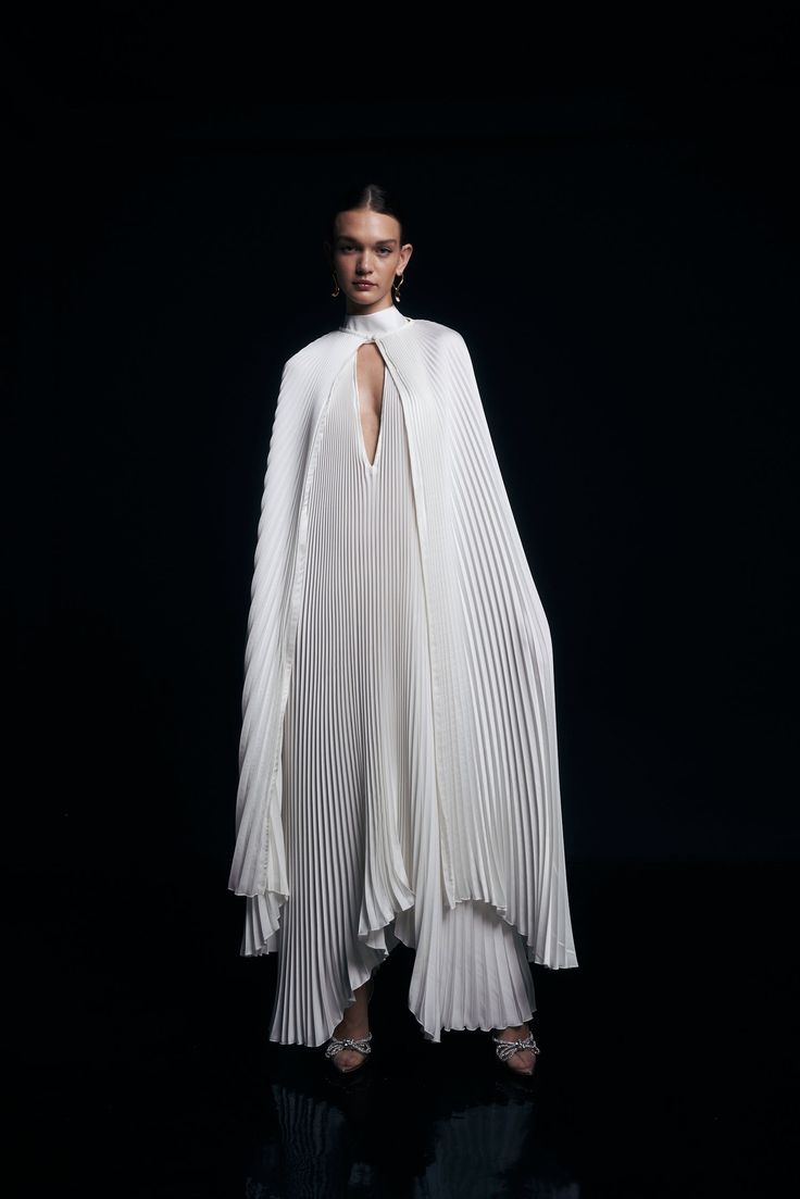 a woman wearing a white pleated cape dress stands with her hands on her hips against a black background. she is wearing minimal jewelry and the cape is partially open to show her neck and collarbone. the dress has pleats throughout and falls to the floor, and the cape falls below her knees.