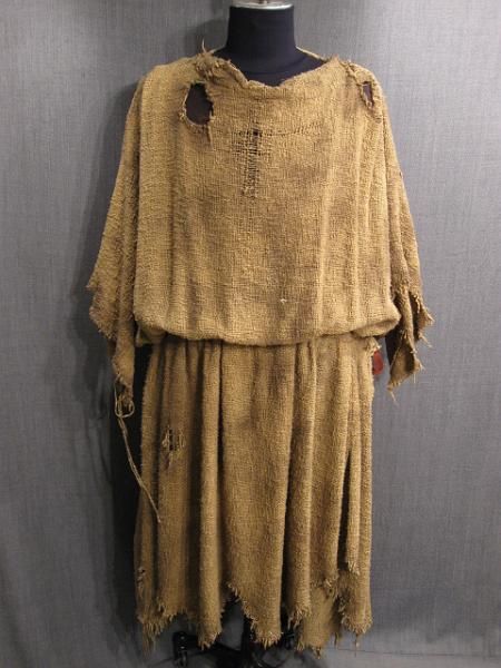 a full shot of a tancolored burlap dress on a mannequin. the dress is old and worn, with several tears and ragged edges. the fabric is rough and has a rustic texture. the dress is styled as a simple, loosefitting tunic over a skirt.  the background is a neutral grey. the overall impression is of an ancient or medievalstyle garment.