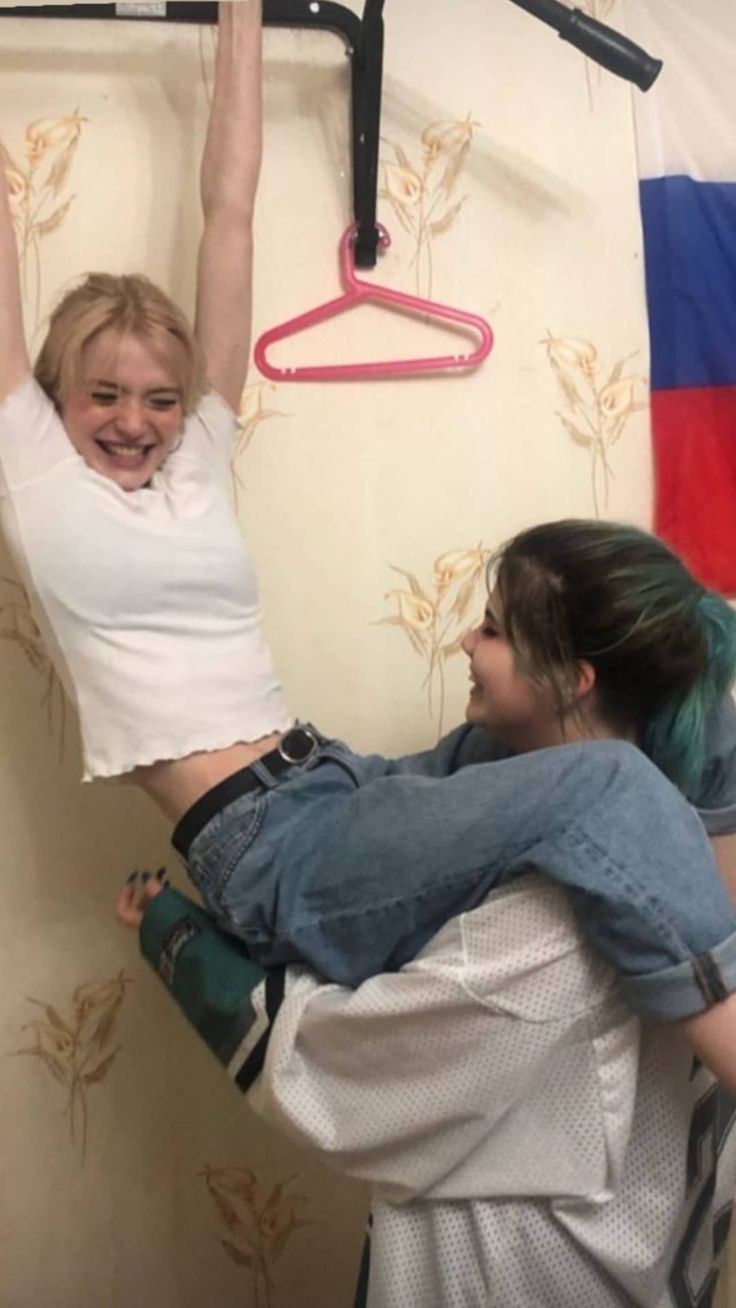 two young girls are hanging out indoors. they are laughing and smiling at each other. one is holding the other upside down. they are both dressed casually in jeans and a white shirt.