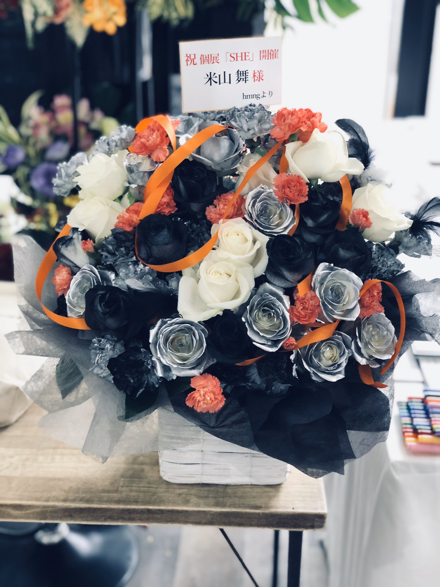 a vibrant floral arrangement featuring black, silver, and white roses accented with orange and coral carnations. an orange ribbon adds a pop of color and a handwritten card in japanese suggests the bouquet is a congratulatory gift. the arrangement sits on a wooden table with a blurred background hinting at an indoor setting.