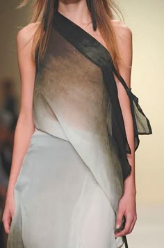a woman is posing in a sheer, draping dress with a scarf that goes over one shoulder, and the fabric drapes across her body. she is on a runway. she is wearing makeup, and her hair is long, straight, and pulled to one side. her expression is neutral.
