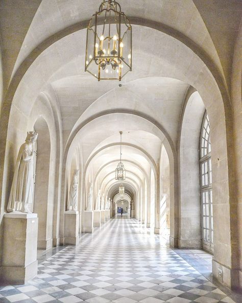 a long corridor inside a palace featuring a black and white checkered floor, white walls and arched ceilings. there are statues on pedestals lining each side of the hallway, as well as large windows along the right. ornate chandeliers hang from the ceiling, providing illumination. there's a single person standing at the end of the hallway.  the perspective lines draw the viewer's eye towards the vanishing point at the end of the hallway.