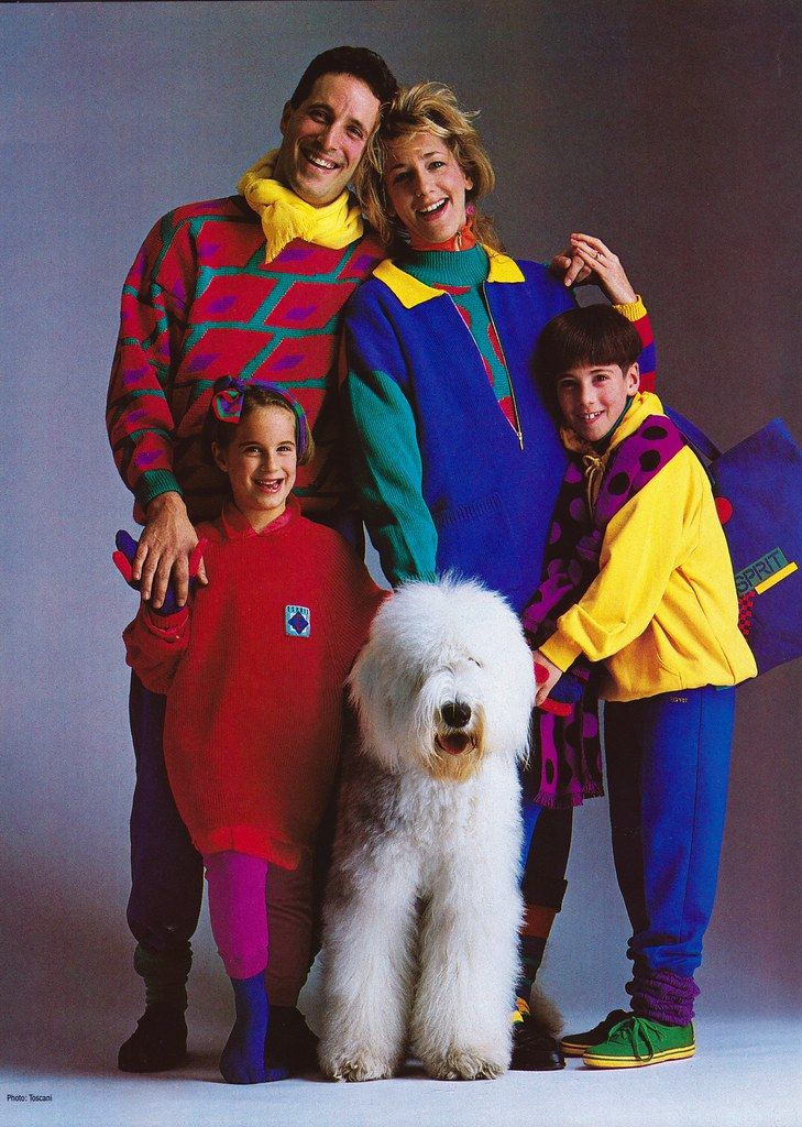 a family of four, including a large, fluffy old english sheepdog, is posing for a photograph. they are all wearing brightly colored, matching or coordinating outfits in a distinct 1980s style. the father is in a red and teal patterned sweater with a yellow scarf. the mother is in a blue sweater with green accents and a yellow collar. the daughter is in a red sweatshirt and pink tights, while the son is wearing a yellow hoodie with black polka dots, blue trousers, and green and yellow sneakers. the dog is sitting in the center of the group. the photograph has a vintage feel and is likely an advertisement or catalog image from the 1980s. the son is holding a blue tote bag that says "esprit" on it.