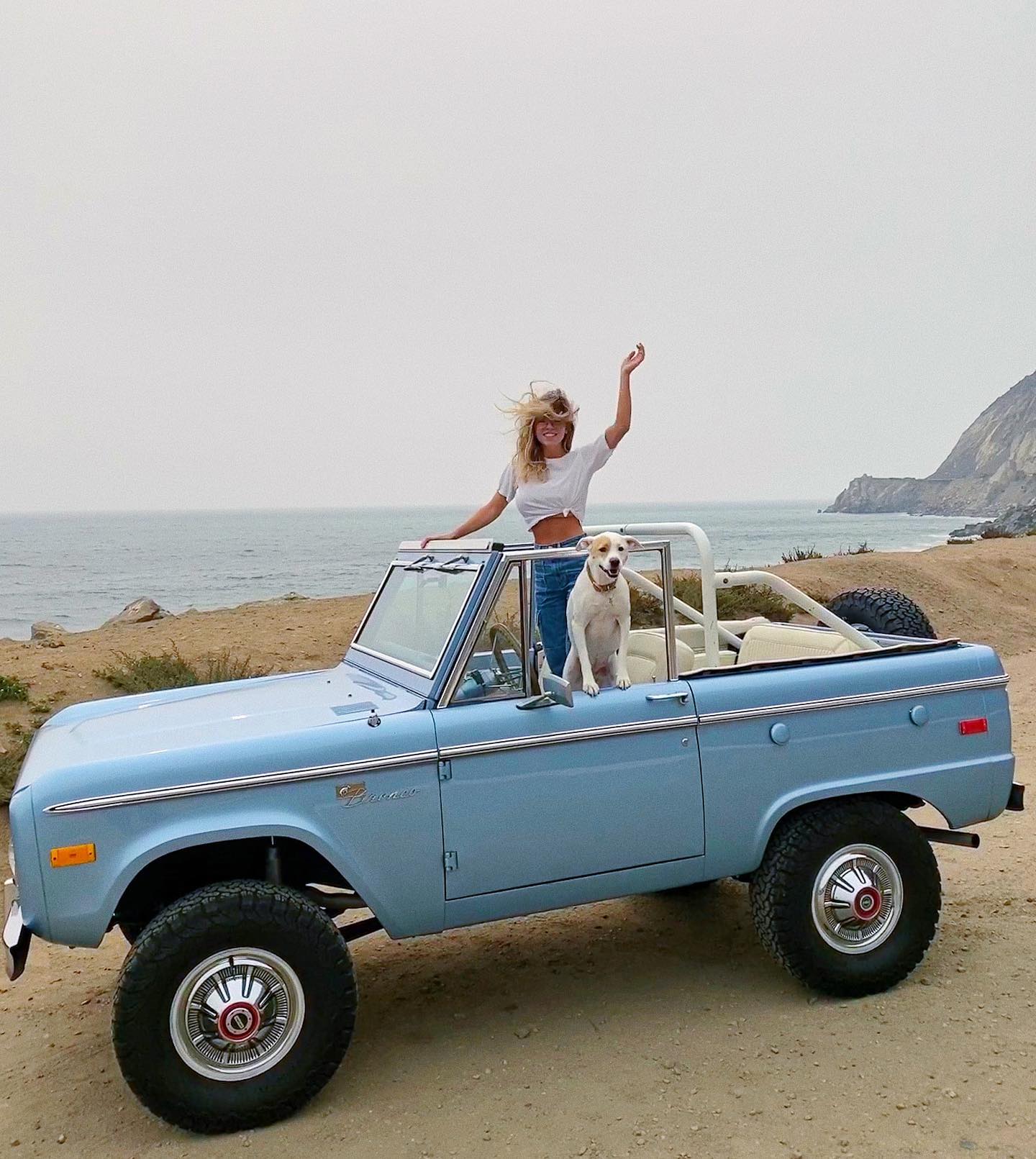 the image shows a light blue vintage ford bronco parked on a sandy beach near the ocean. a young woman with long blonde hair is standing on the back of the vehicle, smiling and waving her arm.  a small white dog is sitting beside her. the background features a clear sky and a scenic coastline. the overall feel is one of summer, freedom, and carefree adventure.