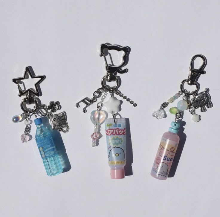 the image shows three keychains, each with unique charms attached. the first keychain has a star clasp and includes a mini blue bottle charm, a butterfly, and several beads. the second keychain features a bearshaped clasp and includes a charm resembling a tube of lotion with a cartoon character on it, along with music note, star, heart shaped bead, and various beads. the third keychain has a standard clasp and includes a mini pink sunscreen bottle charm, an animal charm, and multiple beads. all three keychains have a silver finish.