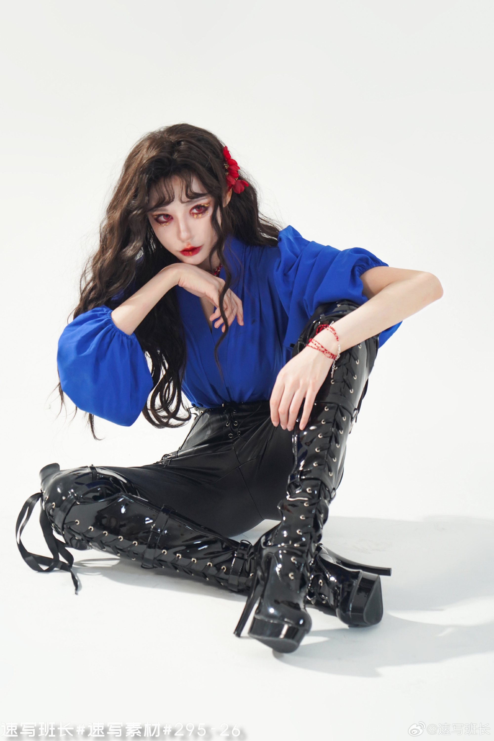 a woman with long dark hair is posing in a studio setting. she is wearing a blue blouse, black patent leather pants, and black laceup platform boots. she has red flowers in her hair and red eye makeup. the background is white.