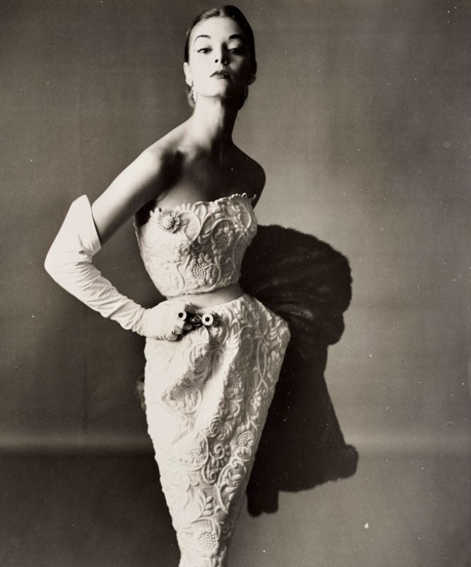 a vintage black and white studio shot of a slender woman in an elegant, embroidered strapless dress and long gloves, posing with style and glamour, against a neutral background with a dark shadow.
