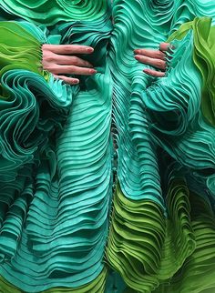 a closeup image of a woman's dress made entirely of ruffled fabric. the dress is a gradient of green shades, starting with a teal green at the top and fading to a lighter green at the bottom. the ruffles are layered and create a wavelike effect, giving the dress a very textural and luxurious appearance. the image is shot from the front, showing the intricate details of the fabric.