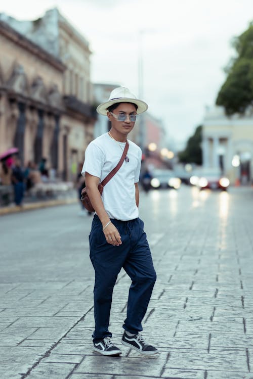 a young man is walking down a city street. he is wearing a white tshirt, dark blue pants, and sneakers. he is also wearing a straw hat and sunglasses. he has a crossbody bag slung across his body. the street is cobblestone. the background is slightly blurred, showing other buildings and people. the overall mood is casual and relaxed.