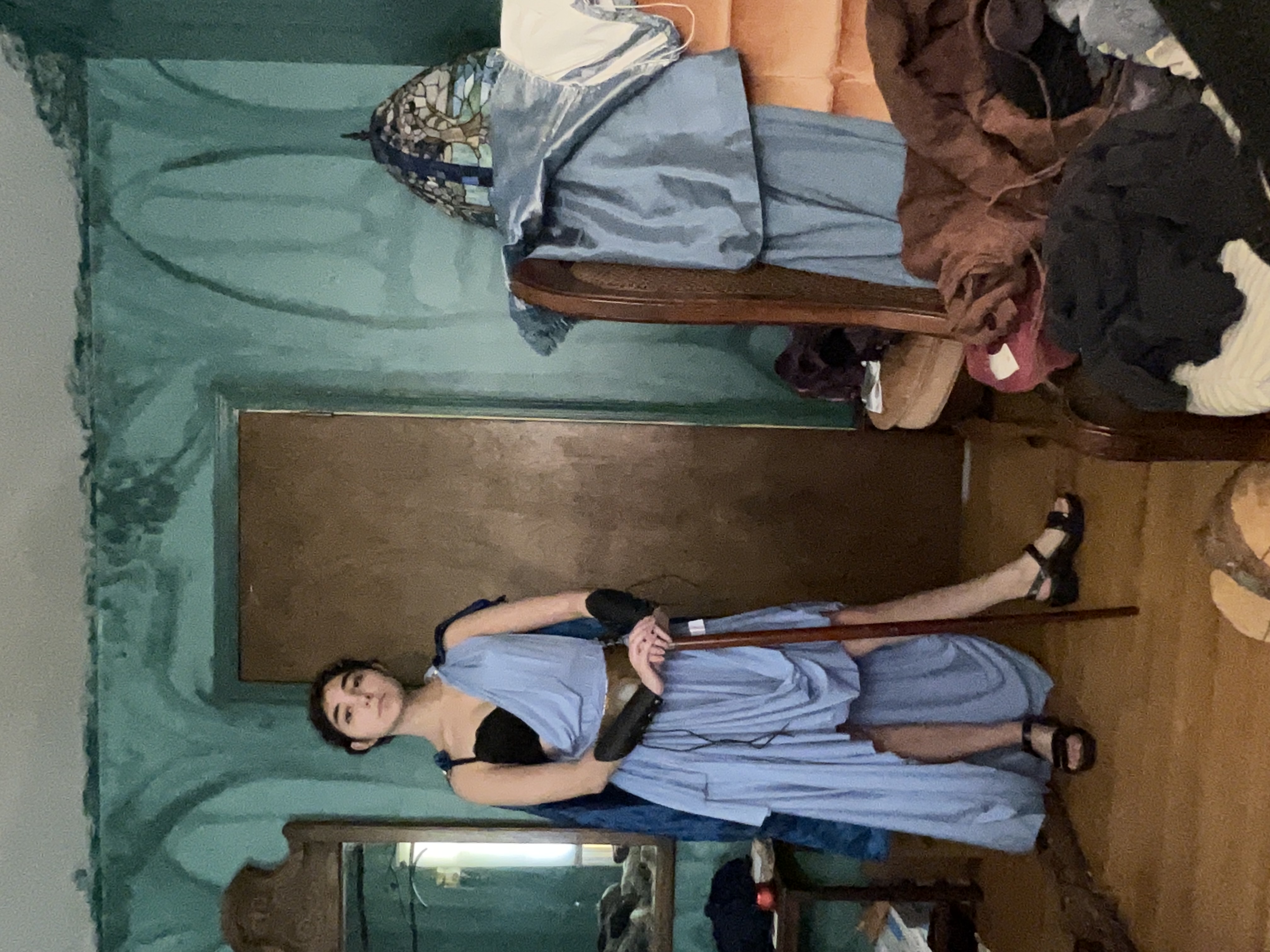 the image shows a young woman dressed in a blue, highlow grecianstyle cosplay costume. she's wearing sandals and holding a wooden cane or walking stick.  the setting is a messy room with tealpainted walls, vintage furniture including an armchair piled with clothes and a mirror. the overall aesthetic suggests a historical or fantasy setting, potentially inspired by the 1920s or 1930s. the room is cluttered, with clothes and other items strewn around, creating a slightly chaotic atmosphere that contrasts with the woman's poised pose.