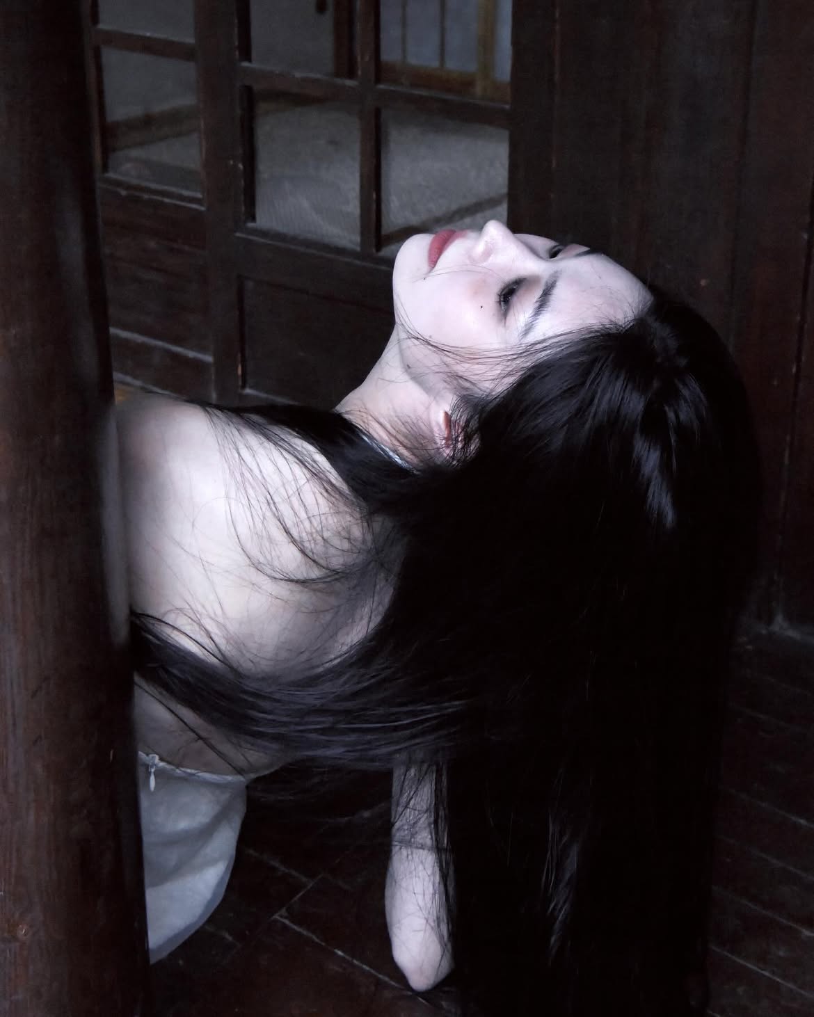 an eerie image of a woman with long, dark hair and pale skin, likely of asian descent, positioned within a wooden structure. her head is tilted back, and she has red lipstick. the overall mood is dark and ghostly.