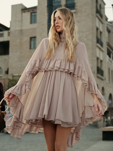 a woman with blonde hair wearing a beige dress with ruffles stands outdoors in an urban setting. the background features a building, adding to the city vibe. she has on jewelry and rings.