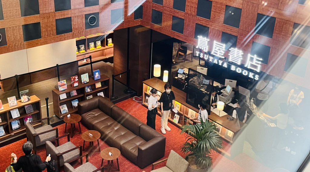 a bird's eye view of a tsutaya bookstore in japan. the bookstore has a modern design with red carpets and wooden shelves. there are several people browsing the shelves and checking out at the counter. the bookstore is welllit with artificial light.