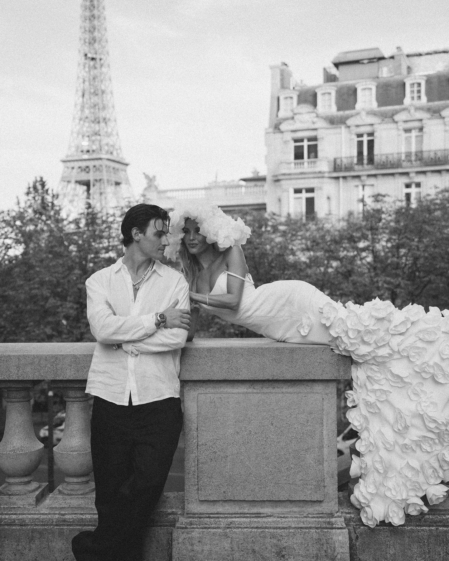 a black and white photo of a couple in paris. the woman is wearing a white dress with a large flower embellishment. the man is wearing a white shirt and black pants. they are both looking at each other and smiling. the eiffel tower is in the background.